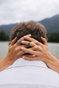 Eine Nahaufnahme der Hände einer Person mit lackierten Nägeln und Ringen, die sanft hinter dem Kopf einer Person verschränkt sind, im Freien in der Nähe von Wasser mit Bergen und bewölktem Himmel im Hintergrund.