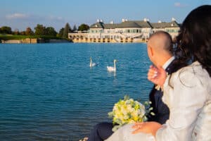 Eine Braut und ein Bräutigam sitzen an einem ruhigen See, halten einen Blumenstrauß in der Hand und schauen auf zwei Schwäne, die zusammen schwimmen, mit einem großen eleganten Gebäude im Hintergrund unter einem klaren Himmel.
