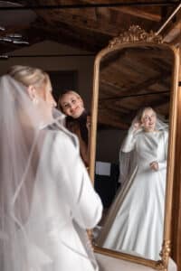 Eine Braut in weißem Kleid und Schleier lächelt, während sie einen Ohrring anlegt und in einen großen, verzierten Spiegel blickt. Eine andere Frau steht hinter ihr, ebenfalls lächelnd, in einem warm beleuchteten Raum mit einer Holzdecke.