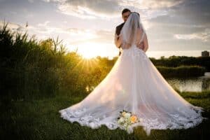Eine Braut und ein Bräutigam stehen bei Sonnenuntergang im Freien dicht beieinander, die Braut in einem langen Spitzenkleid mit Schleier. Das Sonnenlicht leuchtet hinter ihnen und hebt die Schleppe ihres Kleides hervor. Ein Blumenstrauß liegt zu ihren Füßen auf dem Gras in der Nähe eines Teiches und hohen Grases.