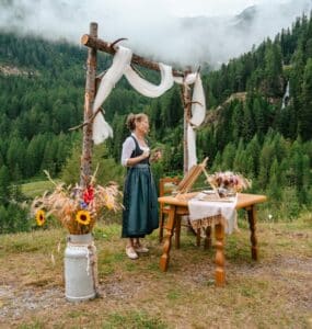 Eine Frau in traditioneller Kleidung steht an einem rustikalen, mit weißem Stoff bespannten Holzbogen neben einem Blumentisch in einer grünen Berglandschaft mit Bäumen und Nebelschwaden.