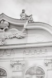 Eine weiße, verschnörkelte Gebäudefassade mit einem geflügelten Emblem und einer sitzenden Statue einer gewandeten Figur über der Dachlinie, mit komplizierten architektonischen Details und einem bewölkten Himmel im Hintergrund.