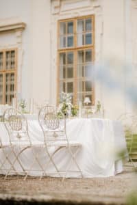 Ein eleganter Tisch im Freien ist mit einem weißen Tischtuch, zarten Blumenarrangements und weißen Stühlen vor den großen Fenstern eines stattlichen Gebäudes gedeckt. Die Szene wirkt hell und luftig und lässt auf eine formelle Veranstaltung schließen.