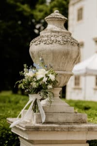 Eine große, verschnörkelte Steinurne steht auf einem Sockel im Freien, geschmückt mit einem Strauß weißer und hellblauer Blumen, die mit einem weißen Band gebunden sind. Im Hintergrund sind ein Gebäude und üppiges Grün zu sehen.