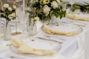 Elegant gedeckter Tisch mit Tellern aus Klarglas, gelben Servietten, Speisekarten, Kristallgläsern und floralen Tafelaufsätzen mit weißen und gelben Blumen, alles auf einem weißen Tischtuch.