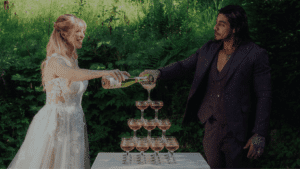 Eine Braut und ein Bräutigam stehen im Freien und schenken Champagner in eine Pyramide von Gläsern auf einem Tisch ein, im Hintergrund Grünzeug und Sonnenlicht.