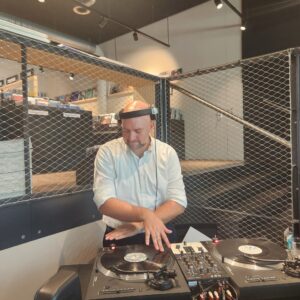 Ein glatzköpfiger Mann im weißen Hemd legt mit Vinyl-Plattenspielern hinter einem Maschendrahtzaun auf und konzentriert sich auf das Abmischen von Musik in einer modernen Innenumgebung mit Regalen im Hintergrund.