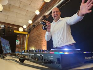 Ein lächelnder Mann in einem weißen Hemd steht hinter einem DJ-Equipment, hält Kopfhörer in einer Hand und streckt den anderen Arm in Richtung Kamera. Im Hintergrund sind helle Lichter und eine Backsteinmauer zu sehen.