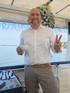 Ein lächelnder Mann mit Glatze und weißem Hemd hält eine weiße Tasse in der Hand und zeigt ein Friedenszeichen auf einem Bootsdeck, mit einem DJ-Pult neben ihm und einem ruhigen Gewässer und Häusern im Hintergrund.