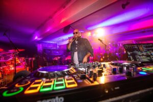 Ein DJ mit Sonnenbrille und Jeansjacke steht auf der Bühne, hält ein Mikrofon in der Hand, ist von bunten Lichtern und einer Menschenmenge umgeben und hat einen DJ-Controller und eine Mischanlage im Vordergrund.