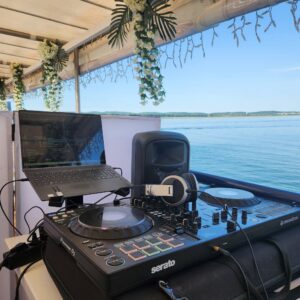 Ein DJ-Setup mit Laptop, Kopfhörern und Mischpult sitzt auf einem mit weißen Blumen und Grünpflanzen geschmückten Boot mit Blick auf das ruhige blaue Wasser unter dem klaren Himmel.