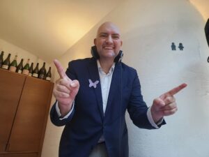 Ein lächelnder glatzköpfiger Mann im Anzug mit einer lila Schmetterlingsnadel und Kopfhörern um den Hals zeigt mit beiden Händen nach oben. Hinter ihm sind Weinflaschen auf einem Schrank vor einer weißen Wand angeordnet.