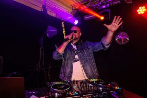 Ein DJ mit Sonnenbrille und Jeansjacke hält ein Mikrofon und hebt eine Hand, während er auf der Bühne auftritt, umgeben von bunten Lichtern und DJ-Equipment, in dessen Nähe eine Discokugel hängt.