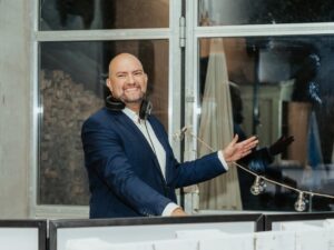 Ein lächelnder Mann im Anzug mit Kopfhörern um den Hals steht hinter einem DJ-Pult und gestikuliert mit einer Hand. Im Hintergrund sind Lichterketten und ein großes Fenster zu sehen.