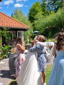 Eine Braut in einem weißen Kleid umarmt einen Gast in einem blauen Blumenkleid auf einer sonnigen Hochzeitsfeier. Zwei Frauen in pastellfarbenen Kleidern stehen in der Nähe, eine hält sich vor Überraschung oder Rührung den Mund zu. Üppiges Grün und blauer Himmel umgeben sie.