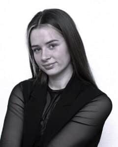 Eine junge Frau mit langem glattem Haar, die ein durchsichtiges schwarzes Oberteil und eine schwarze Weste trägt, lächelt leicht und posiert vor einem schlichten, hellen Hintergrund.