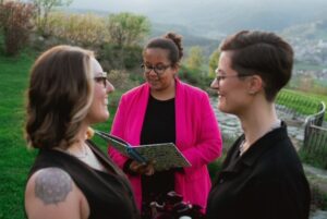 Zwei Frauen stehen sich lächelnd gegenüber, während eine Person in einer leuchtend rosafarbenen Jacke ihre Trauung im Freien mit Bergen und Grün im Hintergrund vollzieht.