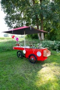 Ein roter fahrbarer Wagen mit einem großen schwarz-roten Regenschirm steht auf einer Wiese in der Nähe von Bäumen. Der Wagen ist mit Luftballons, Flaschen und Blumen geschmückt und hat einen Weidenkorb an der Vorderseite angebracht.