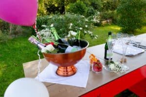 Ein Eiskübel aus Kupfer, gefüllt mit Sektflaschen und weißen Rosen, steht auf einem Tisch im Freien mit Gläsern, einer Flasche und einer Schale mit Erdbeeren. Im Vordergrund sind rosa und weiße Luftballons zu sehen, im Hintergrund ist Grünzeug zu sehen.