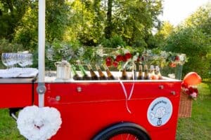 Ein leuchtend roter, blumengeschmückter Barwagen mit Wein- und Sektflaschen, Weingläsern und Eis steht im Freien in einem grünen Park mit Bäumen im Hintergrund.
