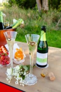 Zwei Sektflöten mit Blasen und Goldringen stehen auf einem Holztisch mit einer Sektflasche, einem Eiskübel, Erdbeeren, Orangenscheiben, weißen Blumen, einem Korken und Grünzeug im Hintergrund.