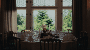 Ein runder Esstisch mit Gläsern, Servietten und einem floralen Tafelaufsatz steht neben großen Fenstern mit Blick auf die Bäume im Freien.