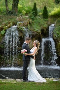 Eine Braut und ein Bräutigam stehen zusammen an einem Teich vor einem kleinen Wasserfall und blicken sich in die Augen. Die Braut hält einen Blumenstrauß in der Hand und trägt ein weißes Kleid, während der Bräutigam einen schwarzen Anzug trägt. Üppiges Grün umgibt sie.