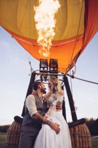 Ein Paar in Hochzeitskleidung umarmt sich und lächelt einander zu, während es im Korb eines Heißluftballons steht und eine große Flamme über ihnen in den Ballon schießt.