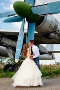Eine Braut und ein Bräutigam stehen dicht beieinander, lächeln und schauen sich an einem sonnigen Tag vor einem großen Flugzeugpropeller an. Die Braut trägt ein weißes Kleid und der Bräutigam ein Hemd mit Krawatte.
