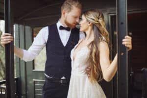Eine Braut in einem weißen Kleid und ein Bräutigam mit Weste und Fliege stehen eng beieinander und blicken sich in die Augen, während sie sich an den Türen eines alten Zugwagens festhalten.