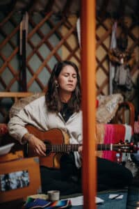 Eine Person mit langen braunen Haaren sitzt im Schneidersitz in einem Raum und spielt mit geschlossenen Augen auf einer Akustikgitarre, umgeben von bunten Textilien und Holzgitterwänden.