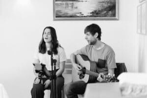 Eine Frau singt, während sie neben einem Mann sitzt, der eine akustische Gitarre spielt. Vor ihnen befinden sich ein Mikrofon und ein Tablet auf einem Ständer. Hinter ihnen hängt ein Gemälde an der Wand. Das Bild ist in schwarz-weiß gehalten.