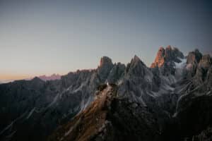 Zerklüftete Berggipfel bei Sonnenaufgang, weiches Licht beleuchtet die felsige Landschaft. Ein schmaler Grat führt auf die Berge zu. In der Mitte steht eine Person, die die dramatische Aussicht unter einem klaren Himmel betrachtet.