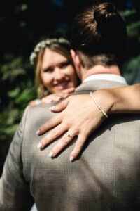 Eine Frau mit einer Blumenkrone umarmt einen Mann im Anzug und lächelt in die Kamera. Ihre Hand, die ein Perlenarmband und einen Verlobungsring trägt, ruht auf seiner Schulter. Der Hintergrund ist im Freien und von der Sonne beschienen.