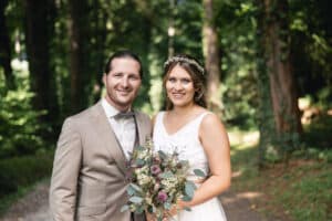 Eine lächelnde Braut und ein lächelnder Bräutigam stehen zusammen in einem sonnenbeschienenen Wald, die Braut hält einen Strauß aus Wildblumen und Grünzeug. Der Bräutigam trägt einen hellen Anzug und eine Fliege, während die Braut ein weißes Kleid und eine Blumenkrone trägt.