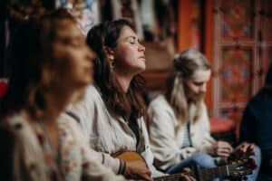 Drei Frauen sitzen in einem Innenraum zusammen; die Frau in der Mitte spielt mit geschlossenen Augen Gitarre und scheint in die Musik vertieft zu sein, während die anderen in der Nähe sitzen, eine unscharf im Vordergrund und eine schreibend oder zeichnend.