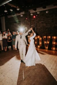 Ein Brautpaar tanzt in einem warm beleuchteten Raum, während die Gäste zuschauen und im Hintergrund große beleuchtete "LOVE"-Buchstaben zu sehen sind. Der Bräutigam nimmt die Braut unter den Arm, und hinter den beiden geht ein Kamerablitz los.