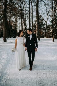 Eine Braut und ein Bräutigam gehen Hand in Hand einen verschneiten Waldweg entlang und lächeln sich dabei an. Die Braut trägt ein weißes Kleid und hält einen Blumenstrauß in der Hand, der Bräutigam trägt einen dunklen Anzug. Kahle Bäume und Schnee umgeben sie.