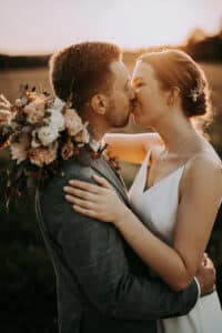Eine Braut und ein Bräutigam küssen sich bei Sonnenuntergang im Freien. Die Braut hält einen Blumenstrauß und trägt ein weißes Kleid, während der Bräutigam einen Anzug trägt. Warmes Sonnenlicht beleuchtet sanft den romantischen Moment.