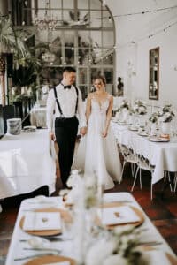 Braut und Bräutigam gehen Hand in Hand durch eine elegant dekorierte Hochzeitsfeier mit weißer Bettwäsche, Blumenschmuck und Lichterketten über dem Kopf.
