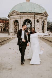 Ein fröhliches Paar in Hochzeitskleidung geht lächelnd und lachend zusammen, während um sie herum Konfetti vor einem Kuppelpavillon im Freien fällt.