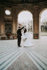 Eine Braut und ein Bräutigam tanzen zusammen unter einem verzierten, offenen Pavillon mit gewölbten Fenstern, umgeben von Winterbäumen und grauem Himmel. Die Braut hält einen Blumenstrauß und lächelt, während der Bräutigam sie auf dem gemusterten Boden herumwirbelt.