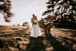 Eine Braut und ein Bräutigam stehen zusammen auf einem sonnenbeschienenen Feld neben einem Baumstumpf, umgeben von hohem Gras und Bäumen, wobei das warme Sonnenlicht lange Schatten hinter sie wirft.