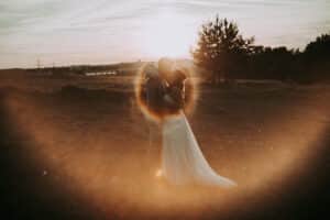Ein Paar in Hochzeitskleidung umarmt und küsst sich in einem Feld bei Sonnenuntergang, umgeben von einem warmen orangefarbenen Streulicht, das einen leuchtenden Ring um sie herum bildet.
