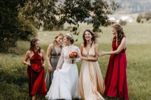 Eine Braut in einem weißen Kleid und vier Brautjungfern in farbenfrohen Kleidern lachen gemeinsam im Freien auf grünem Gras unter einem Baum, mit einer verschwommenen Landschaft im Hintergrund.