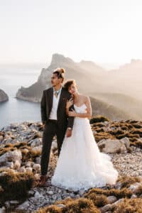 Eine Braut in einem weißen Kleid lehnt sich an einen Bräutigam im Anzug, während sie auf einem felsigen Gelände mit Küstenklippen und dem Meer im Hintergrund steht, das in warmes Sonnenlicht getaucht ist.