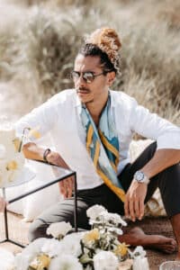 Ein Mann mit lockigem Haar in einem Dutt, der eine Brille, ein weißes Hemd, einen bunten Schal und eine dunkle Hose trägt, sitzt barfuß auf dem Boden neben einem Tisch, umgeben von weißen und gelben Blumen im Freien.