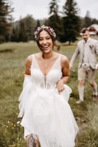 Eine lächelnde Braut in einem weißen Kleid und einer Blumenkrone steht im Freien auf einer Wiese und hält ihren Rock. Ein Mann in einem beigen Anzug und kurzen Hosen geht hinter ihr. Im Hintergrund sind Kiefern und ein rustikales Gebäude zu sehen.