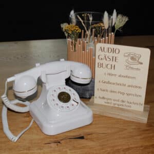 Ein altes weißes Telefon mit Drehscheibe liegt auf einem Holztisch neben einem Schild in deutscher Sprache, das ein Audiogästebuch erklärt, mit einer Vase mit getrockneten Blumen im Hintergrund.