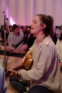 Eine junge Frau sitzt im Schneidersitz, spielt eine akustische Gitarre und singt in ein Mikrofon. Sie ist umgeben von einem sitzenden Publikum in einem sanft beleuchteten Raum mit lila Licht.
